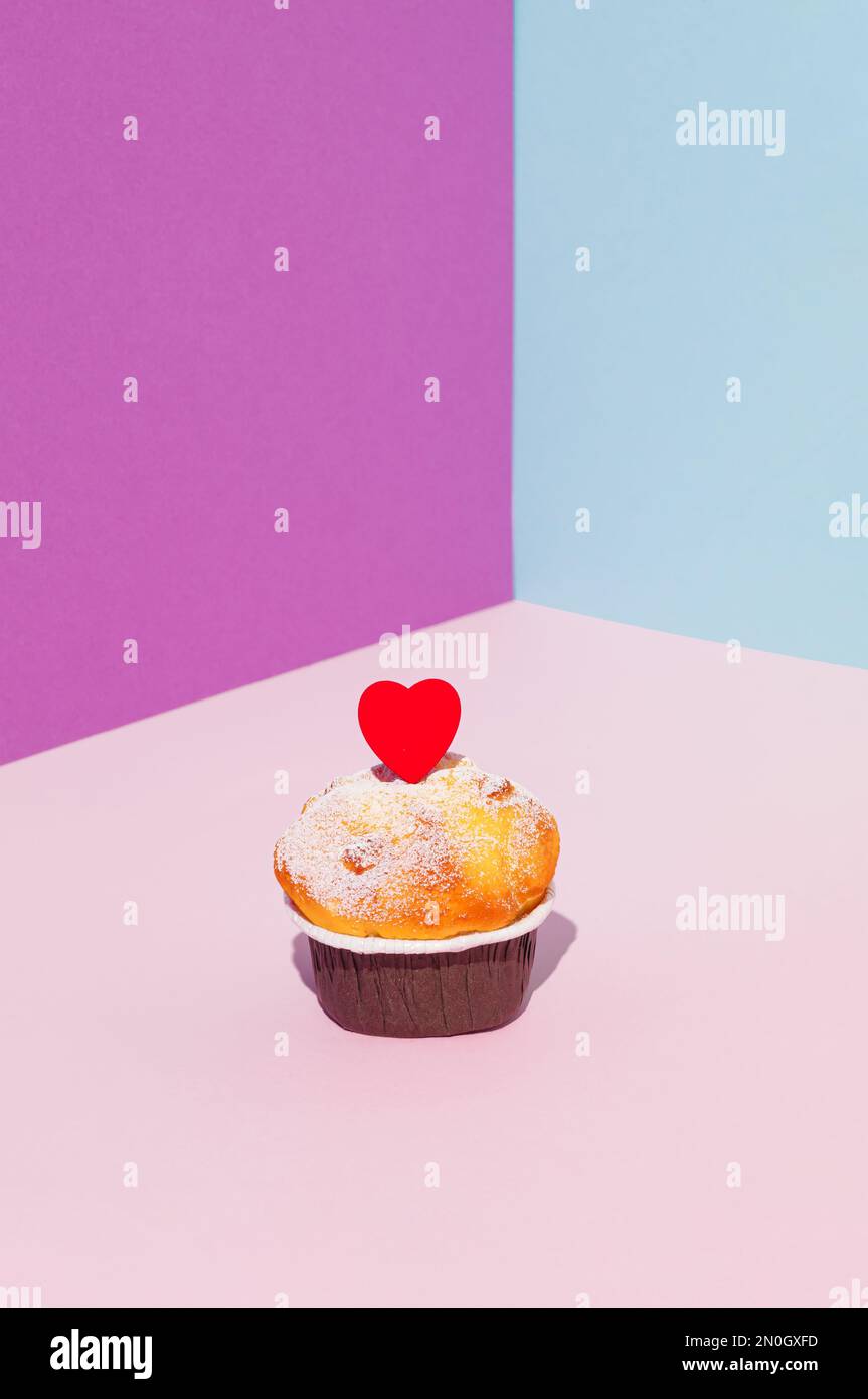 Cupcake mit rotem Holzherz auf violettem, blauem und pinkfarbenem Hintergrund. Minimales Valentinstagskonzept. Stockfoto