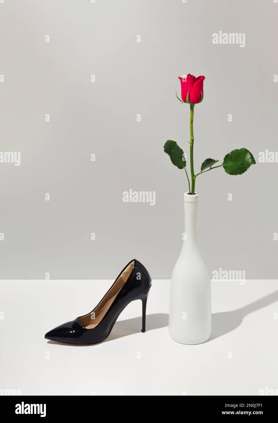 Kreatives Konzept mit Rosenblume, Champagnerflasche und schwarzem Damenschuh. Nachhaltige Modeidee mit weißem und grauem Hintergrund. Minimaler Valentinstag Stockfoto