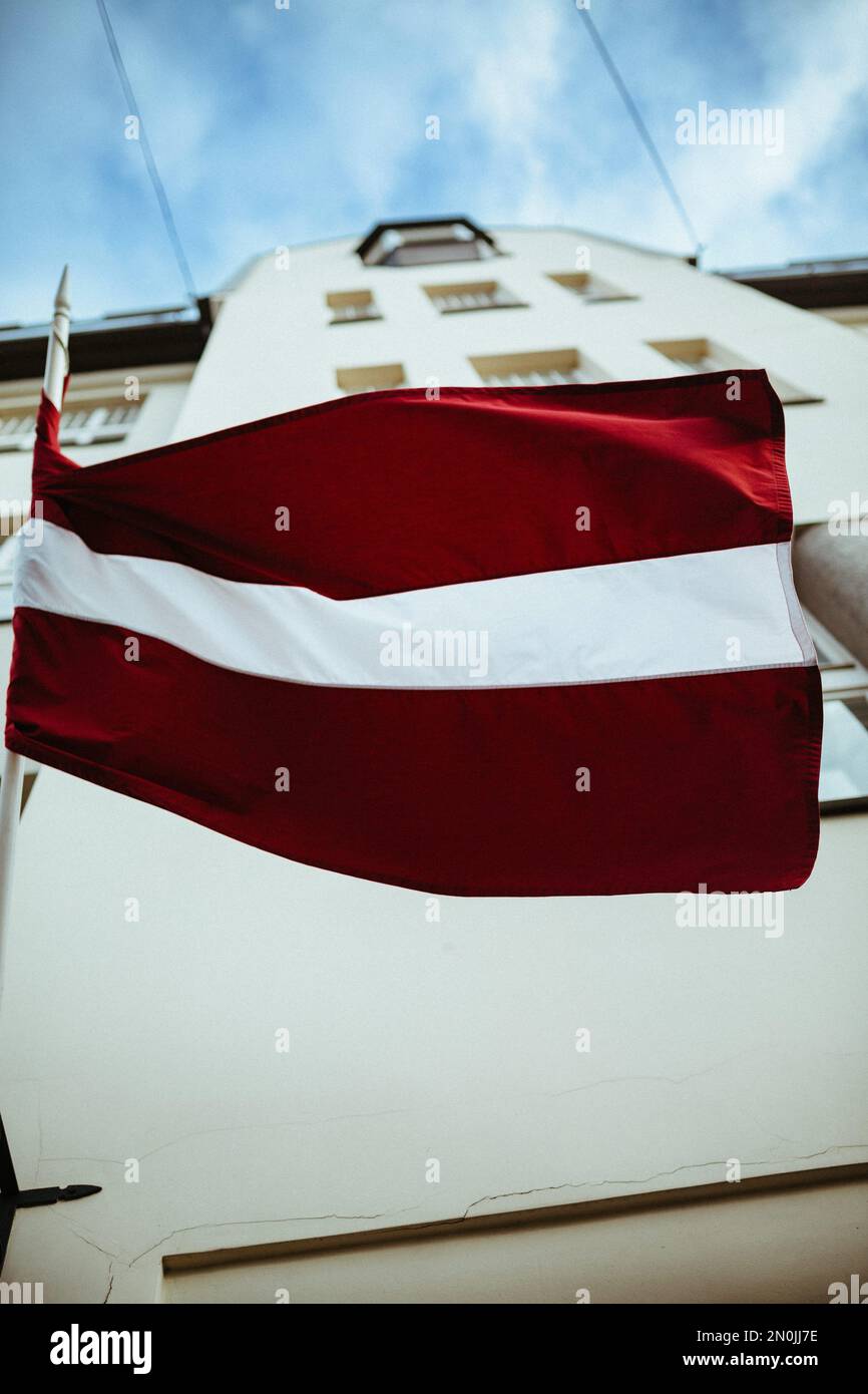 Ein kleiner vertikaler Schuss der lettischen Flagge mit Gebäude vor blauem Himmel Stockfoto