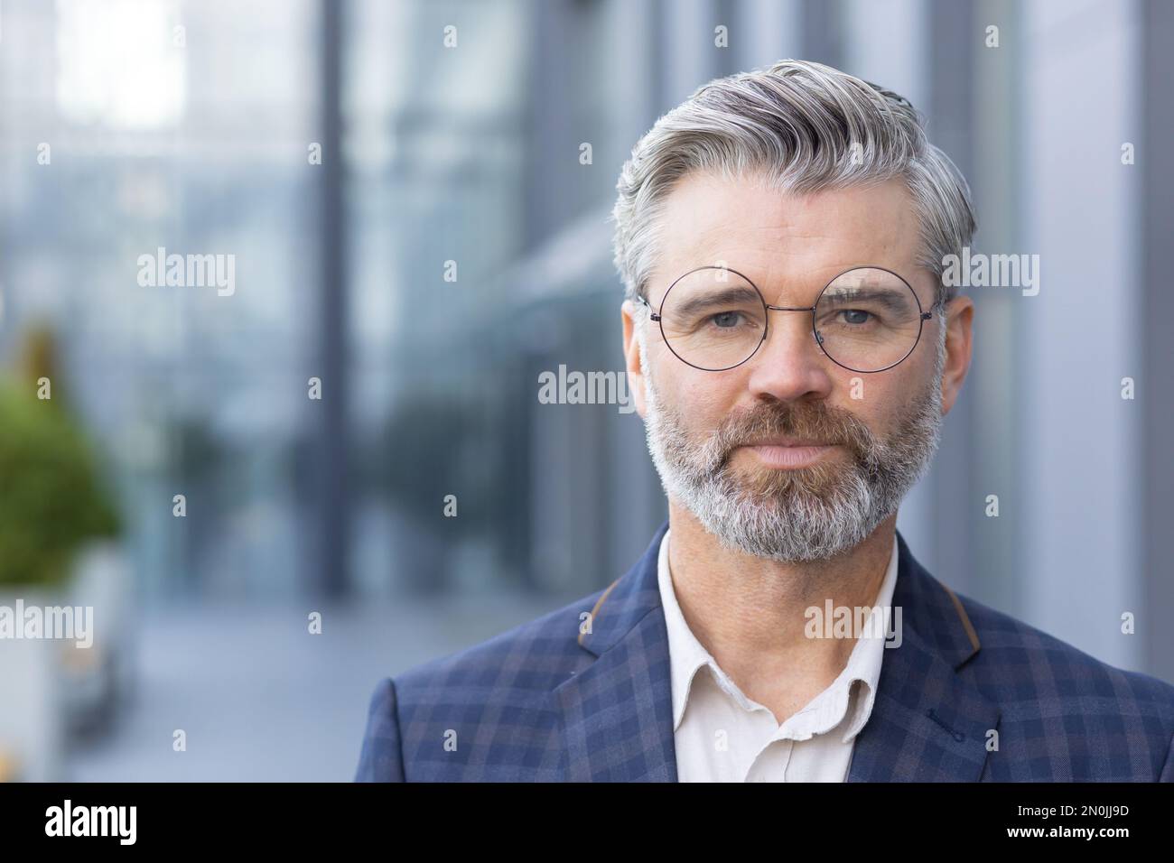 Nahaufnahme eines ernsthaften fokussierten Geschäftsmanns, eines erwachsenen Mannes mit Brille und Business-Anzug, der von außerhalb des Bürogebäudes auf die Kamera schaut. Stockfoto
