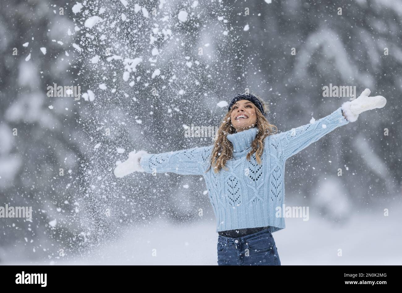 Mit weit geöffneten Armen genießt eine Frau in blauem Pullover, weißen Handschuhen, Schal und Hut an einem verschneiten Wintertag frische Luft. Stockfoto