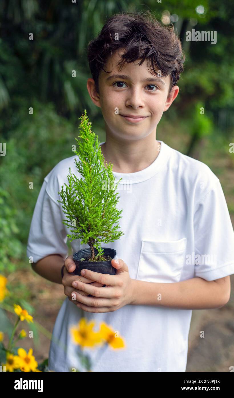 Ein Junge, der einen kleinen Baum in einem Topf hält, in einem Garten steht. Stockfoto