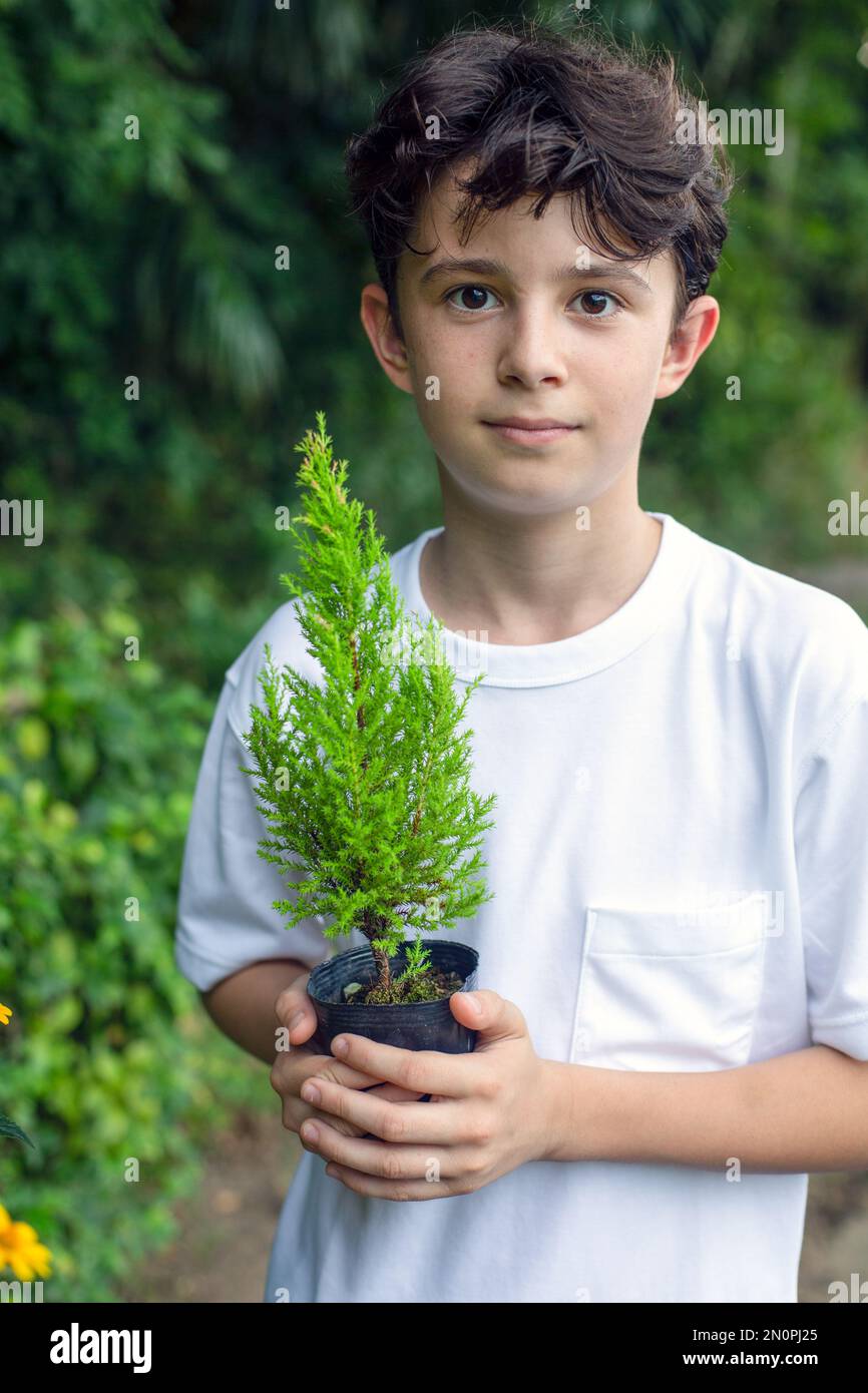 Ein Junge, der einen kleinen Baum in einem Topf hält, in einem Garten steht. Stockfoto