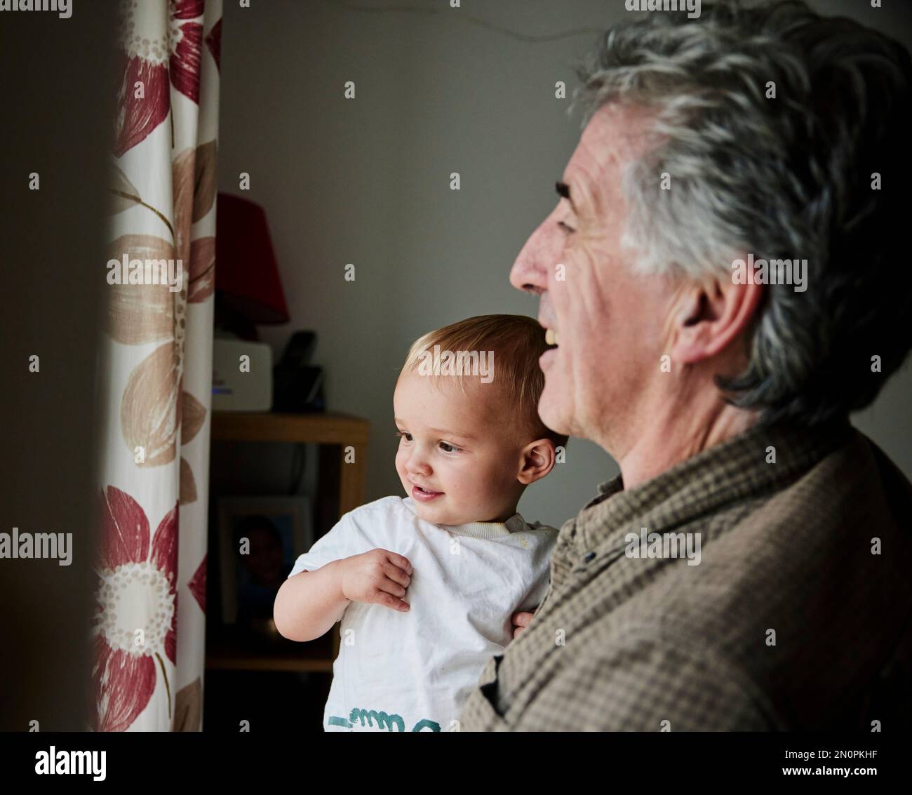Nahaufnahme eines lächelnden Kleinkindes in den Armen eines Großvaters, das aus dem Fenster schaut Stockfoto
