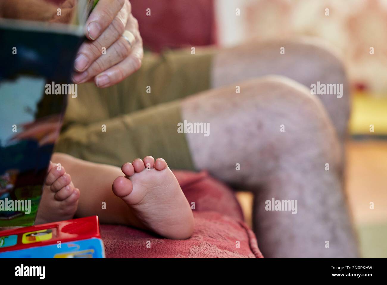 Nahaufnahme des Großvaters, der dem Kleinkind vorliest, Blick auf die barfüßigen Füße und die Knie des Großvaters. Stockfoto