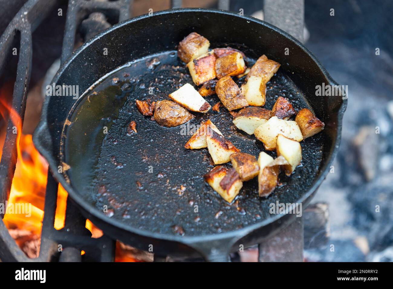 Gusseiserne Pfanne, die gebratene hashbraune Kartoffeln in Speckfett über einem Lagerfeuer kocht Stockfoto