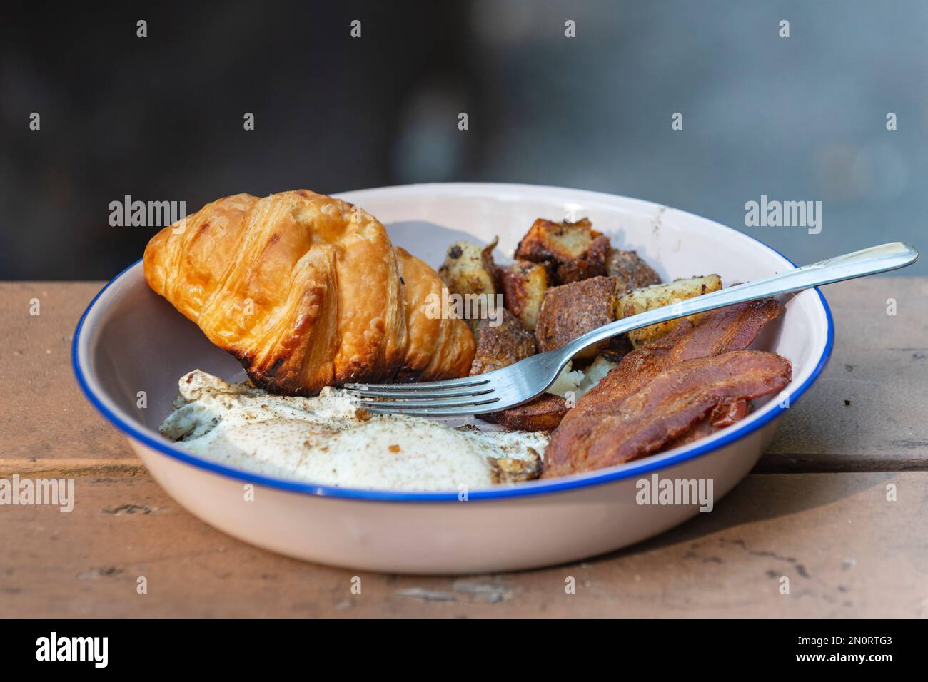 Frühstück mit Eiern, Speck, Kartoffelrösti, Croissant, gekocht in einer gusseisernen Pfanne über einem offenen Feuer. Stockfoto