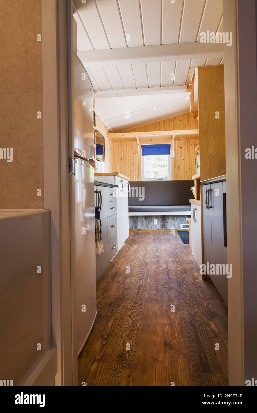 Braun gebeizter Kiefernholzboden und Blick auf Küchenablagen, weiße und graue Melaminschränke und Hauptsitzbereich im 8 x 24 Fuß großen mobilen Mini-Haus. Stockfoto