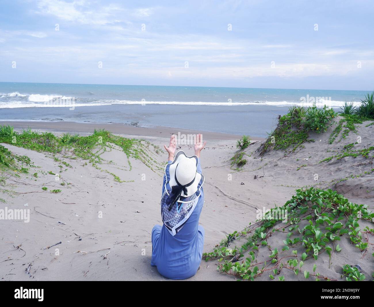 Der Rücken der Frau mit dem Hut am tropischen Strand, die auf den Himmel und das Meer blickt, während sie ihre Hand im Gebet erhebt Stockfoto