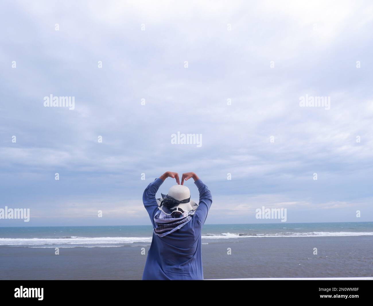 Der Rücken der Frau mit dem Hut am tropischen Strand, die auf den Himmel und das Meer blickt, während ihre Hände Liebe bilden Stockfoto