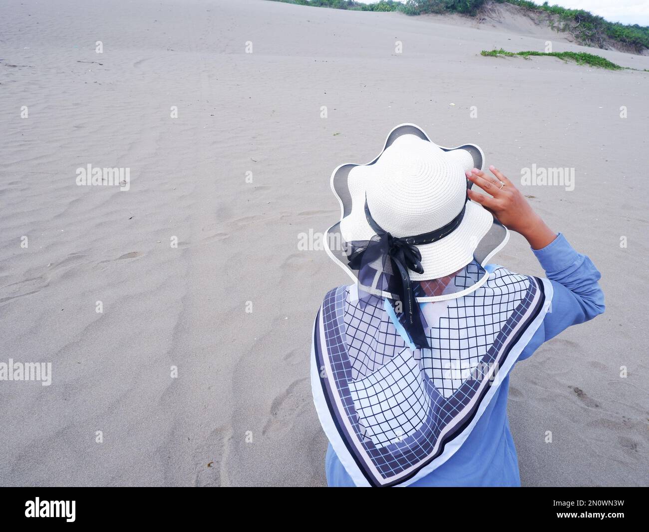 Der Rücken der Frau mit dem Hut am tropischen Strand, die auf dem Strand-Sand saß. Hintergrund von Strand Sand und Himmel. Sandberg Stockfoto