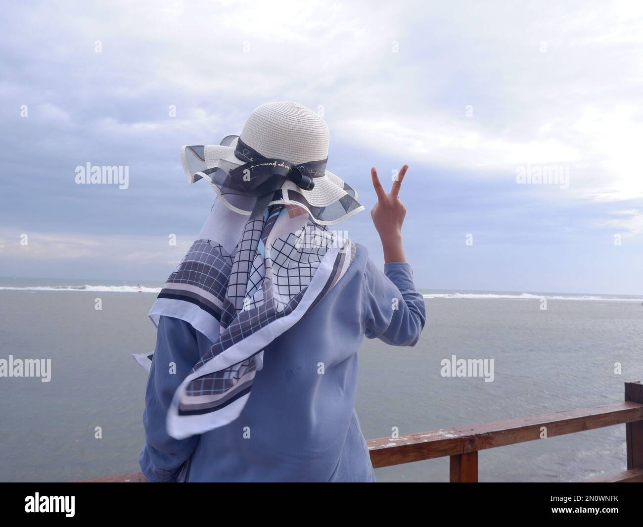 Der Rücken der Frau mit dem Hut am tropischen Strand, die auf den Himmel und das Meer blickt und eine friedliche Geste zeigt. Vertikal. Meereshintergrund Stockfoto