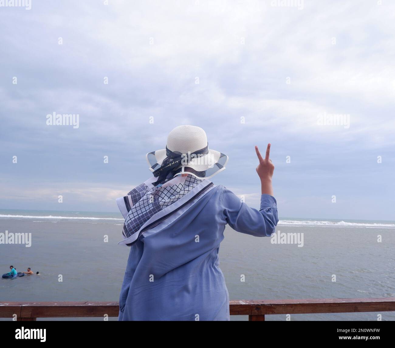 Der Rücken der Frau mit dem Hut am tropischen Strand, die auf den Himmel und das Meer blickt und eine friedliche Geste zeigt. Vertikal. Meereshintergrund Stockfoto