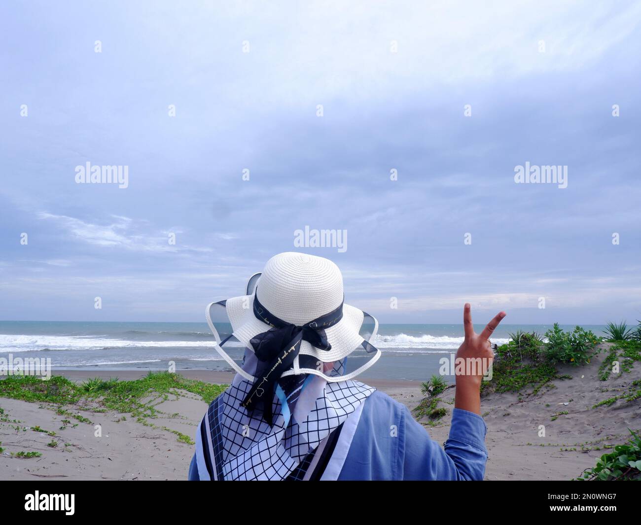 Der Rücken der Frau mit dem Hut am tropischen Strand, die auf den Himmel und das Meer blickt und eine friedliche Geste zeigt. Vertikal. Meereshintergrund Stockfoto