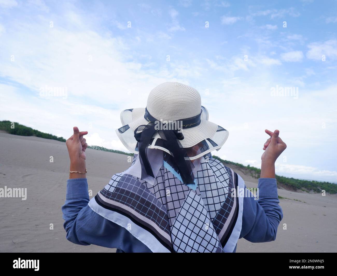 Der Rücken der Frau mit dem Hut am tropischen Strand, die auf das Meer starrte und dann eine Liebe aus der Hand machte. Hintergrund von Strand Sand und Himmel. Be Stockfoto