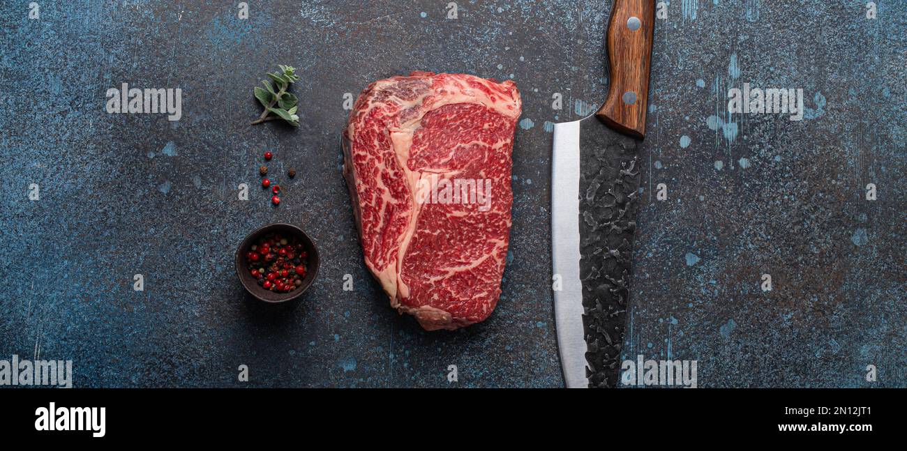 Rohes Rindfleisch Marmorsteak Ribeye auf rustikalem Beton Küchentisch von oben mit großem Messer und Gewürzen, Beefsteak-Konzept Stockfoto