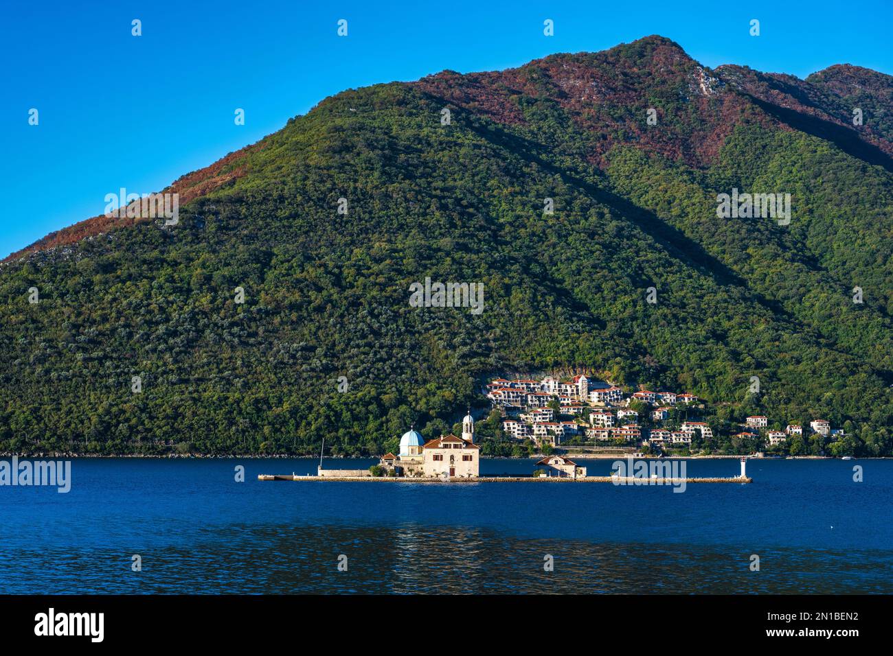 Unsere Frau der Felsen vor der Küste von Perast, mit der kleinen Stadt Kostanjica dahinter, in der Bucht von Kotor in Montenegro Stockfoto