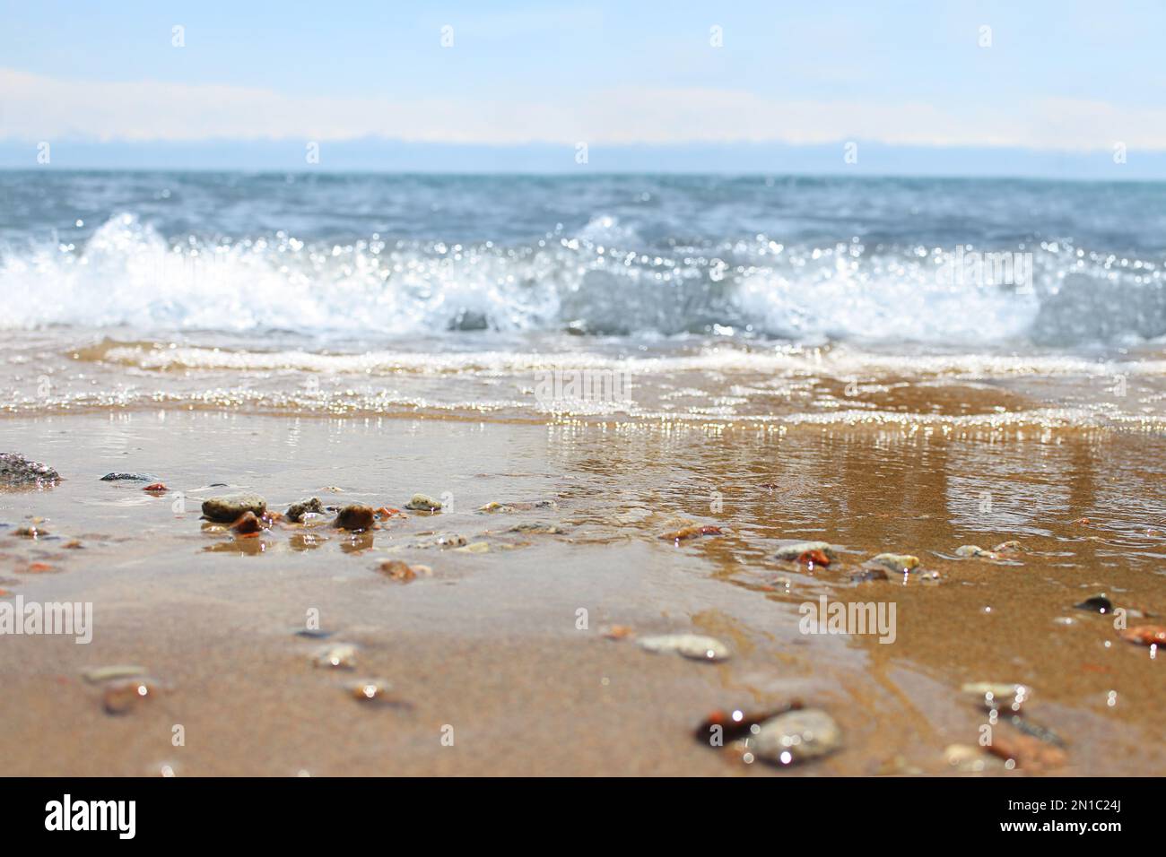 Unschärfe-Strand mit verwischter Welle und nassem Sand mit Kieselsteinen am blauen Himmel. Meer, Meereshintergrund. Outdoor-Urlaub und Reise-Abenteuer-Konzept Stockfoto