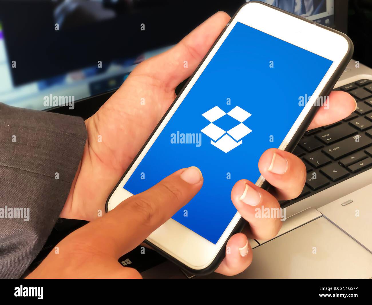 Mann, der die Speicheranwendung dropbox auf Mobilgeräten verwendet, mit Laptop im Hintergrund, redaktioneller technischer Hintergrund Stockfoto