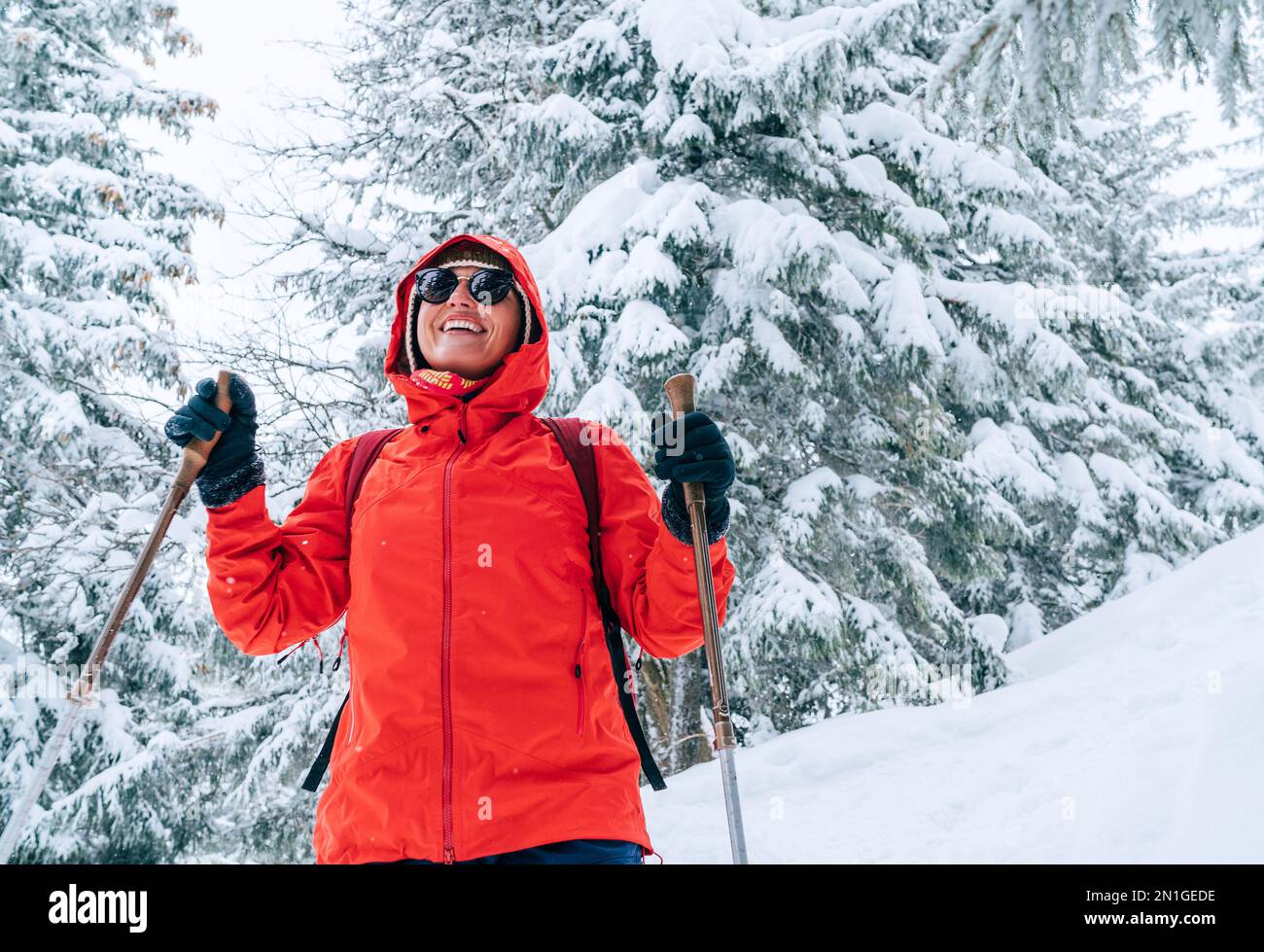 Lächelnde weibliche Trekkerin in roter Sonnenbrille mit Wanderstöcken, die an einem schneebedeckten Hang mit Tannenbäumen und Schnee entlang wandern, niedrige Tatra, SL Stockfoto