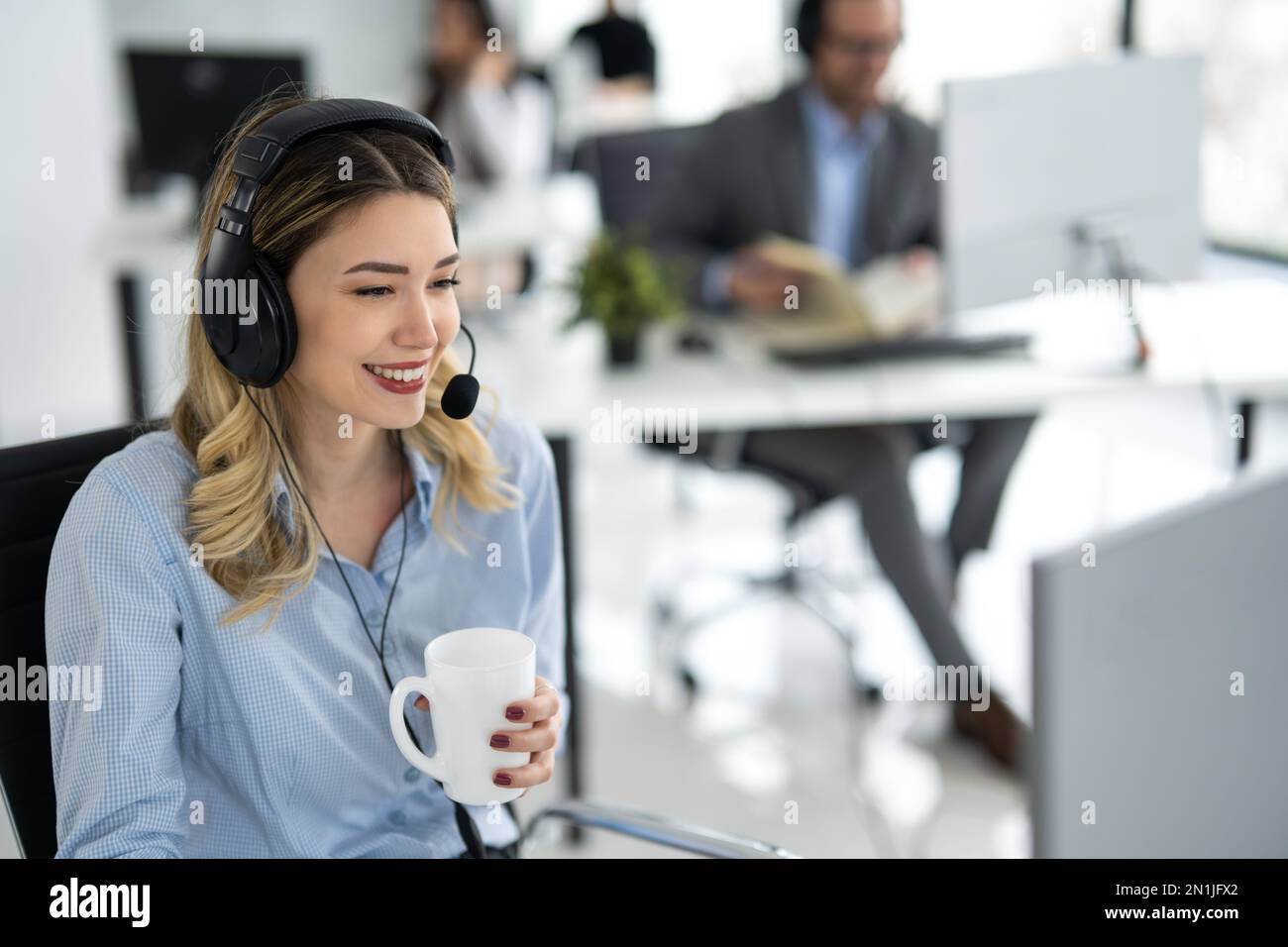Porträt einer schönen jungen Helpline-Mitarbeiterin, die eine Kaffeetasse hält und sich mit einem Kunden im Callcenter-Büro unterhält Stockfoto