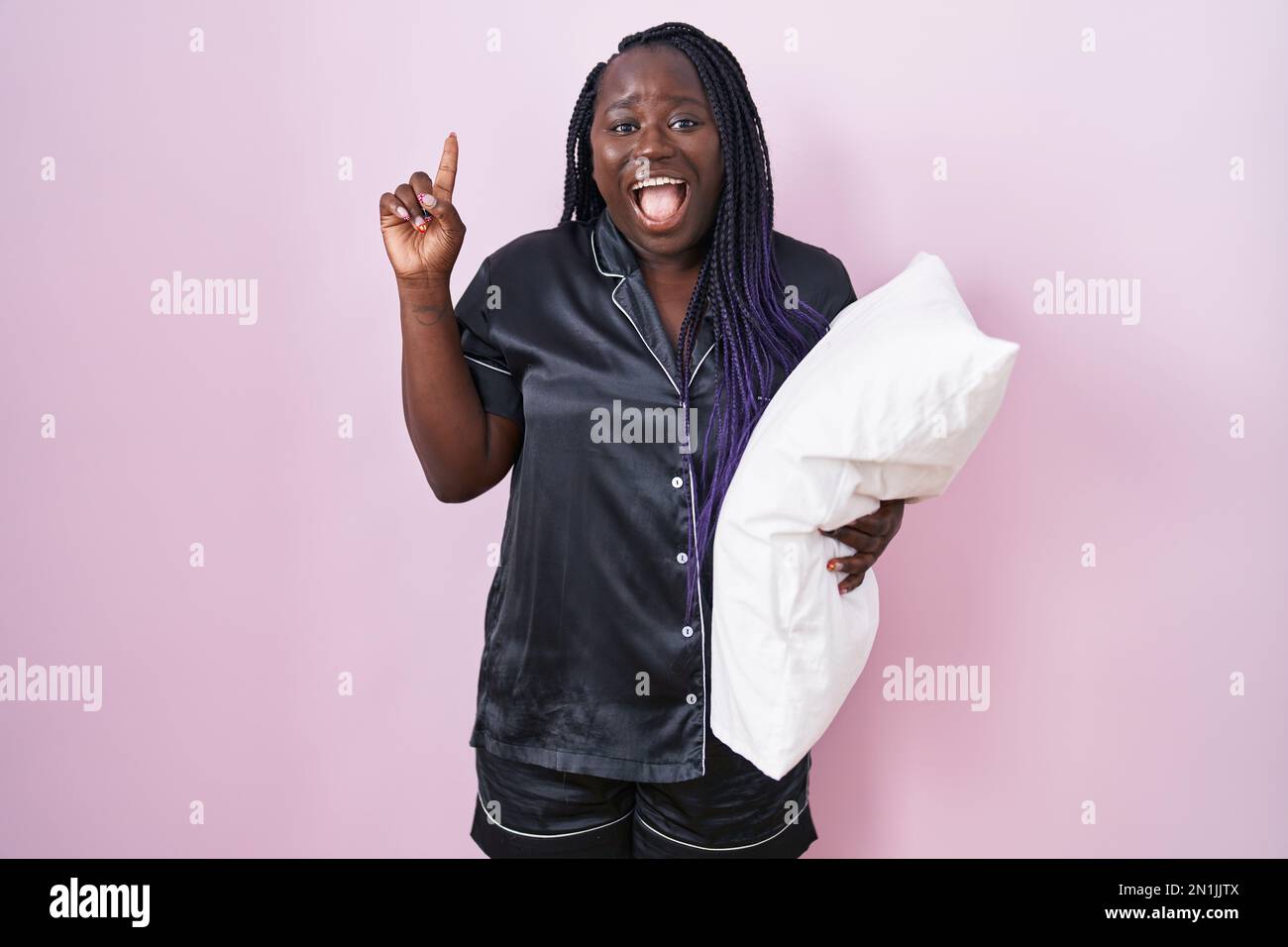 Junge afrikanische Frau, die Pijama-Kissen trägt, lächelt und überrascht und zeigt mit Fingern und erhobenen Armen nach oben. Stockfoto