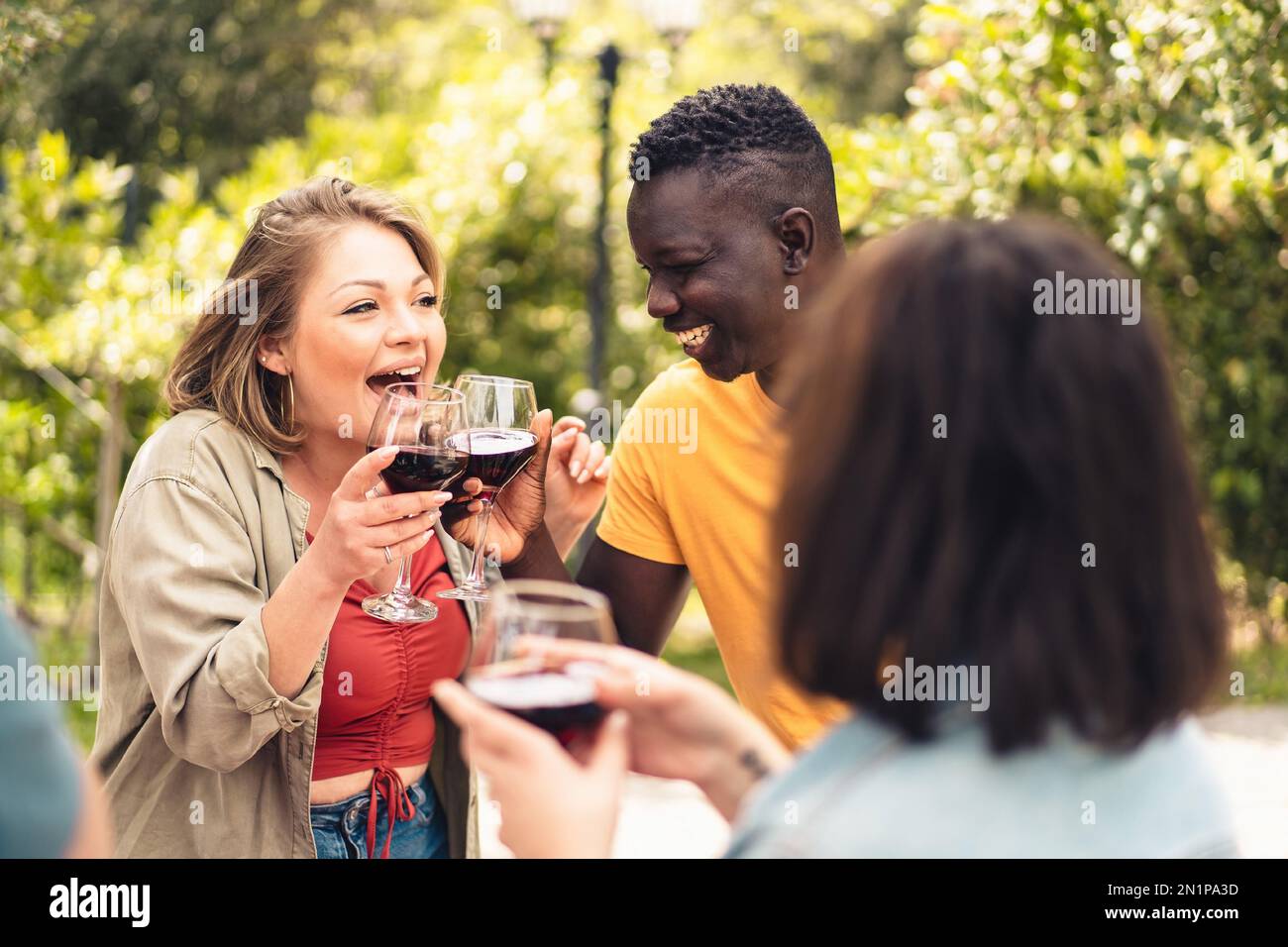 Eine kurvige junge Erwachsene Kaukasierin und ein junger afrikanischer Mann jubeln und lachen, während sie mit Rotwein anstoßen - vielfältige fröhliche Menschen Lebensstil Conc Stockfoto