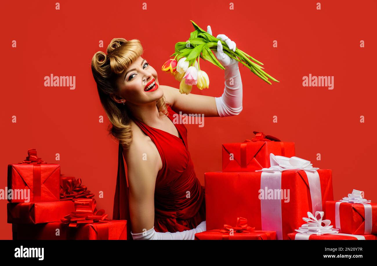 Lächelnde Frau mit Frühlingsblumen. Hübsches blondes Mädchen in rotem Kleid mit Geschenk und einem Strauß Tulpen. Frühlingsblumen und Geschenk. Frau mit Retro Stockfoto