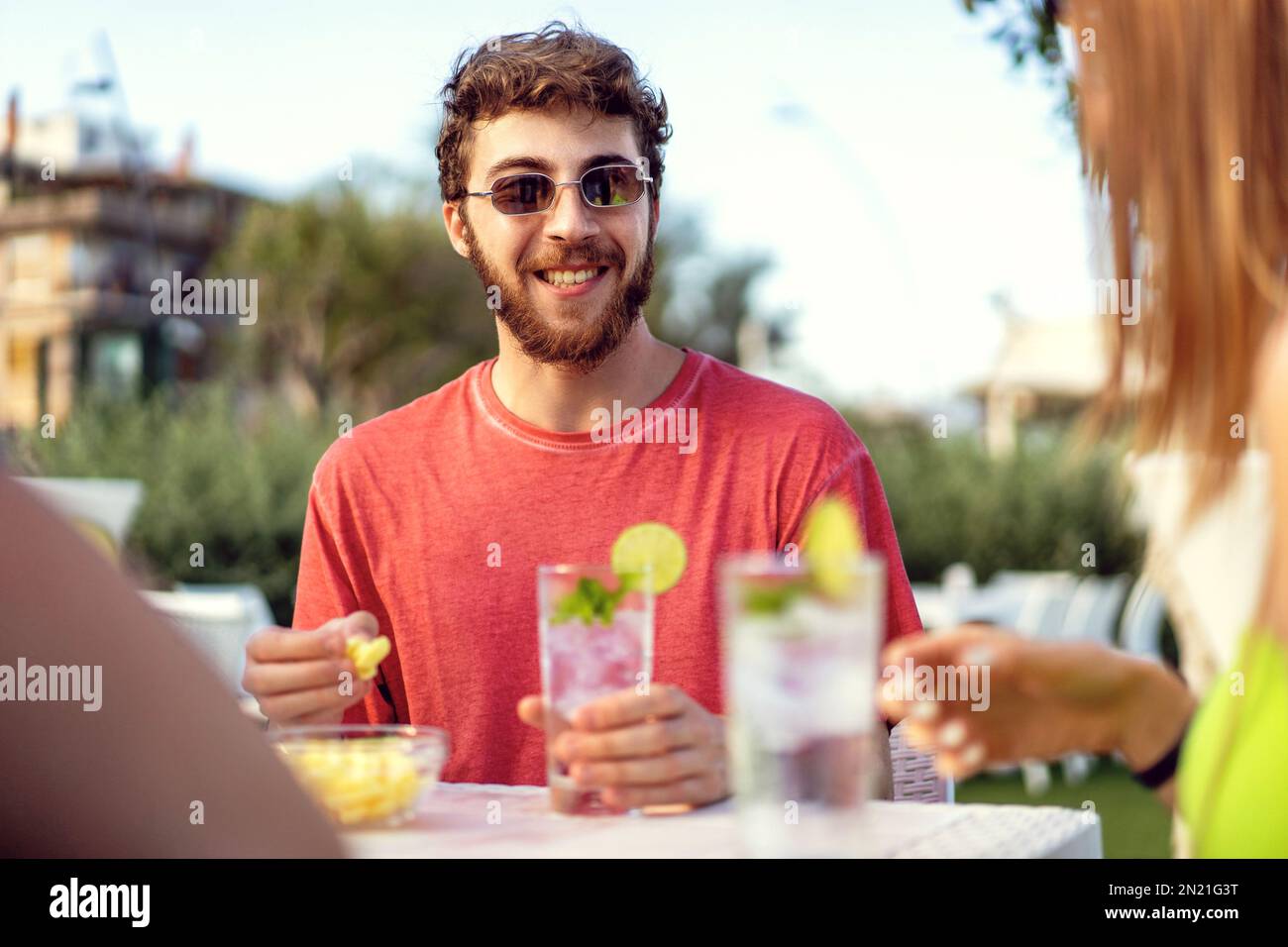 Ein junger Mann sitzt an einem Pub-Tisch im Freien, lächelt und hat eine tolle Zeit mit seinen Freunden (außerhalb der Kamera). Er trägt ein rotes T-Shirt und hält ihn fest Stockfoto