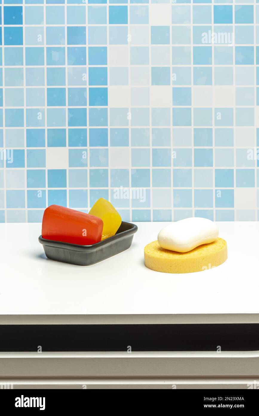 Schüssel aus schwarzem Porzellan mit Seifenriegeln aus Glycerin, einem weißen Seifenriegel und einem gelben Schwamm auf einer weißen Oberfläche zum Händewaschen in einem blau gefliesten Badezimmer Stockfoto