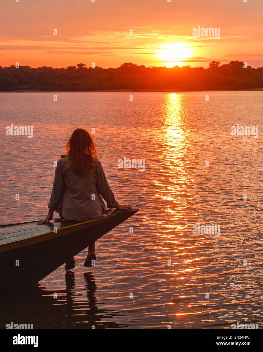 Ein vertikales Bild einer Frau, die auf einem Pier sitzt und die Aussicht auf den Sonnenuntergang genießt Stockfoto