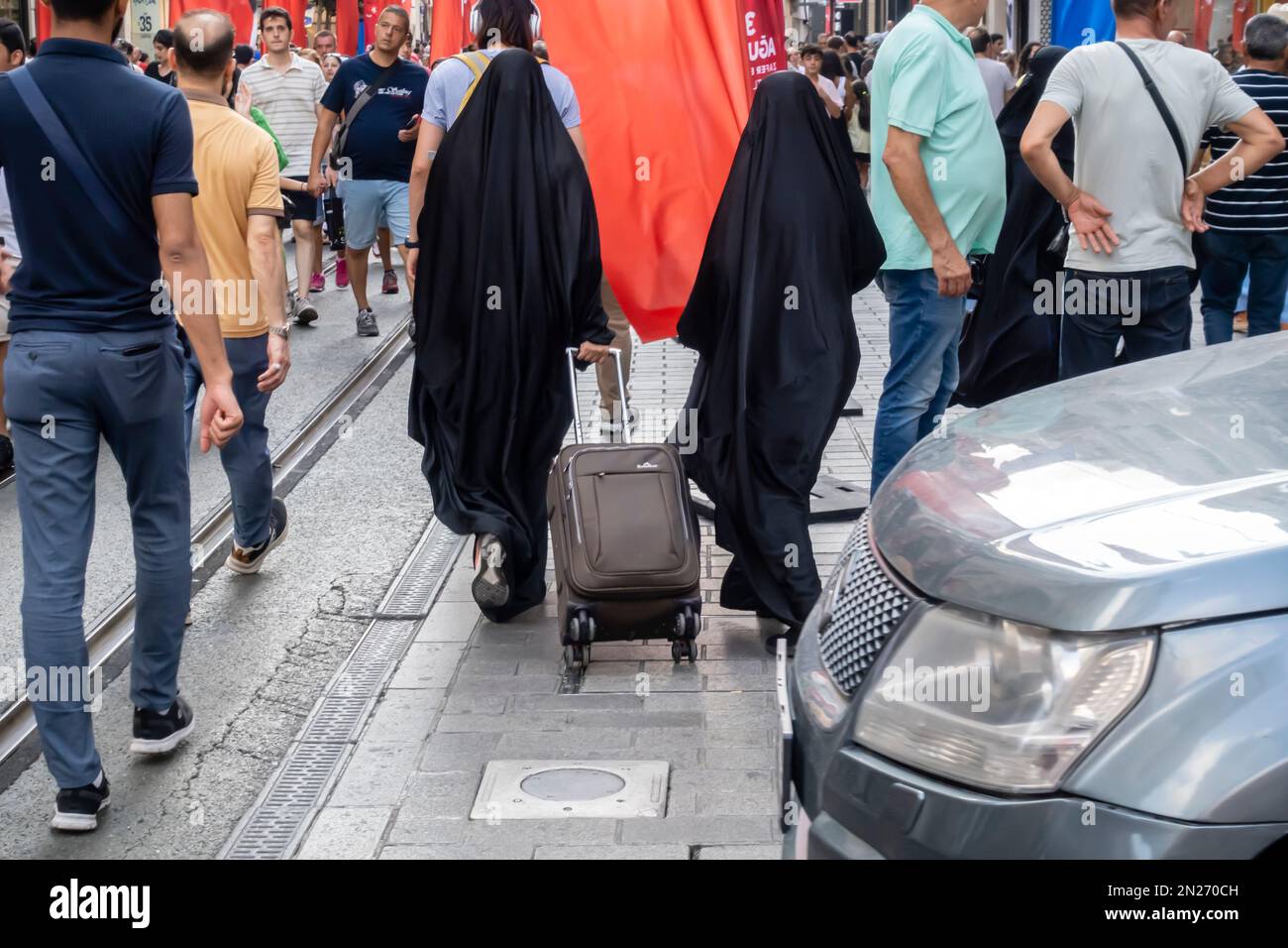 Weibliche Touristen in schwarzen Burqas, Hijabs mit rollendem Gepäck gehen auf der belebten Straße mit multikulturellen Menschenmassen im Zentrum von Istanbul, Türkei Stockfoto