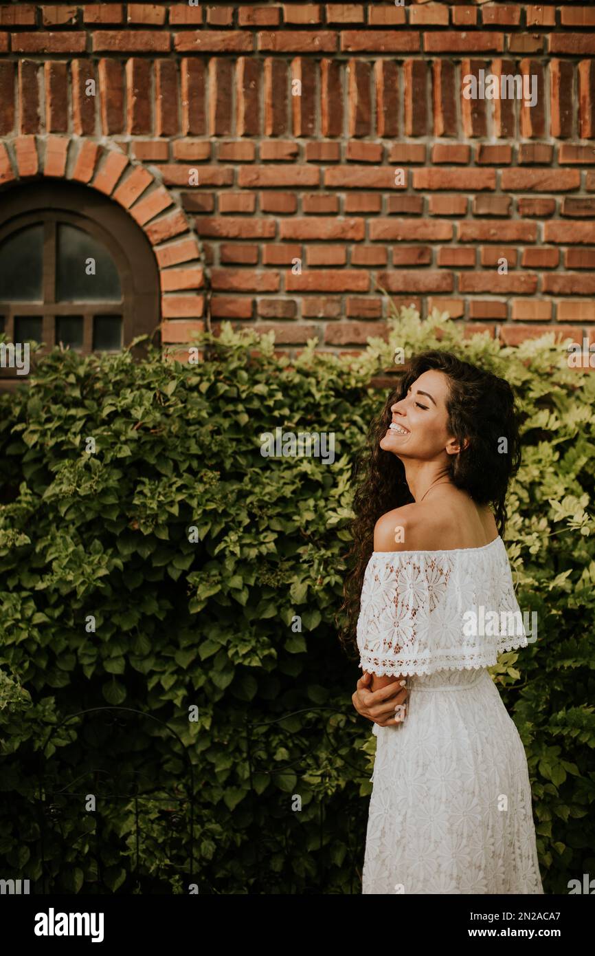 Hübsche junge Frau mit lockigem Haar steht in einem wunderschönen Garten, gekleidet in einem weißen Kleid. Sie sieht entspannt und zufrieden aus Stockfoto