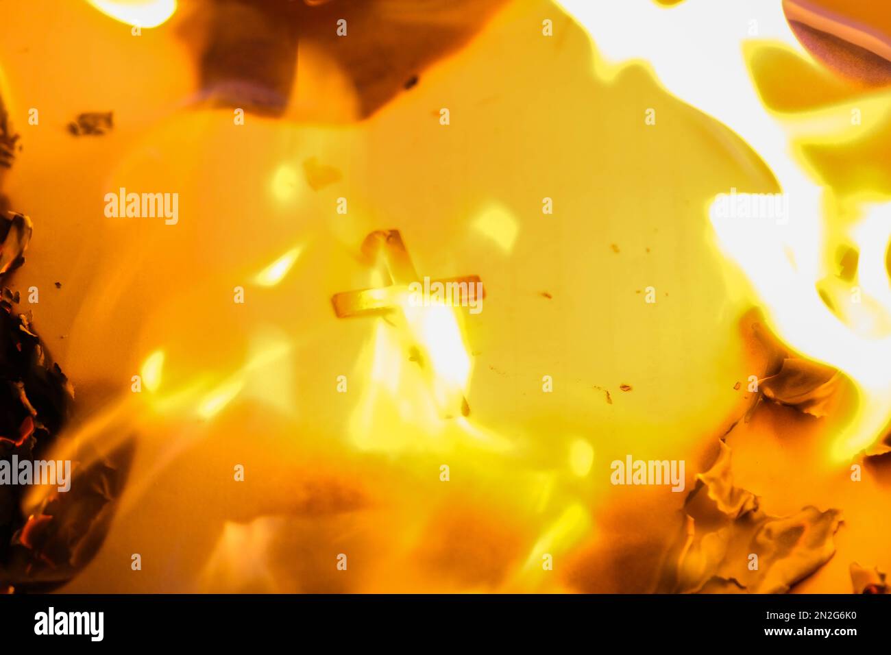 Flammen auf einem brennenden silbernen Kreuz, die in der Nacht glühen. Nahaufnahme brennendes Papier mit silbernem Kreuz in der Mitte auf schwarzem Hintergrund. Stockfoto