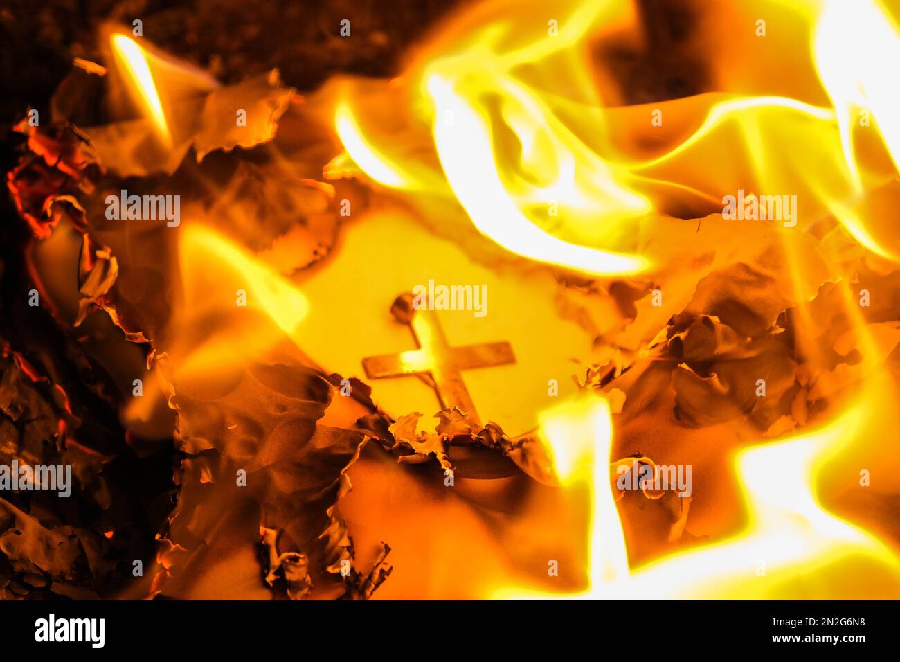 Flammen auf einem brennenden silbernen Kreuz, die in der Nacht glühen. Nahaufnahme brennendes Papier mit silbernem Kreuz in der Mitte auf schwarzem Hintergrund. Stockfoto