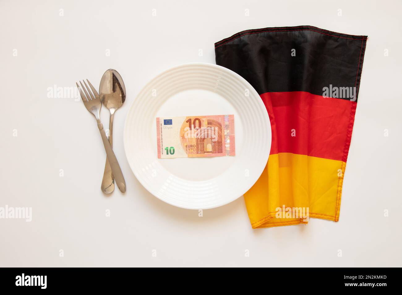 10 Euro liegen auf einem Teller neben einem Löffel und einer Gabel und der Flagge Deutschlands auf weißem Hintergrund, Finanzen und Wirtschaft in Deutschland, Wirtschaft Stockfoto