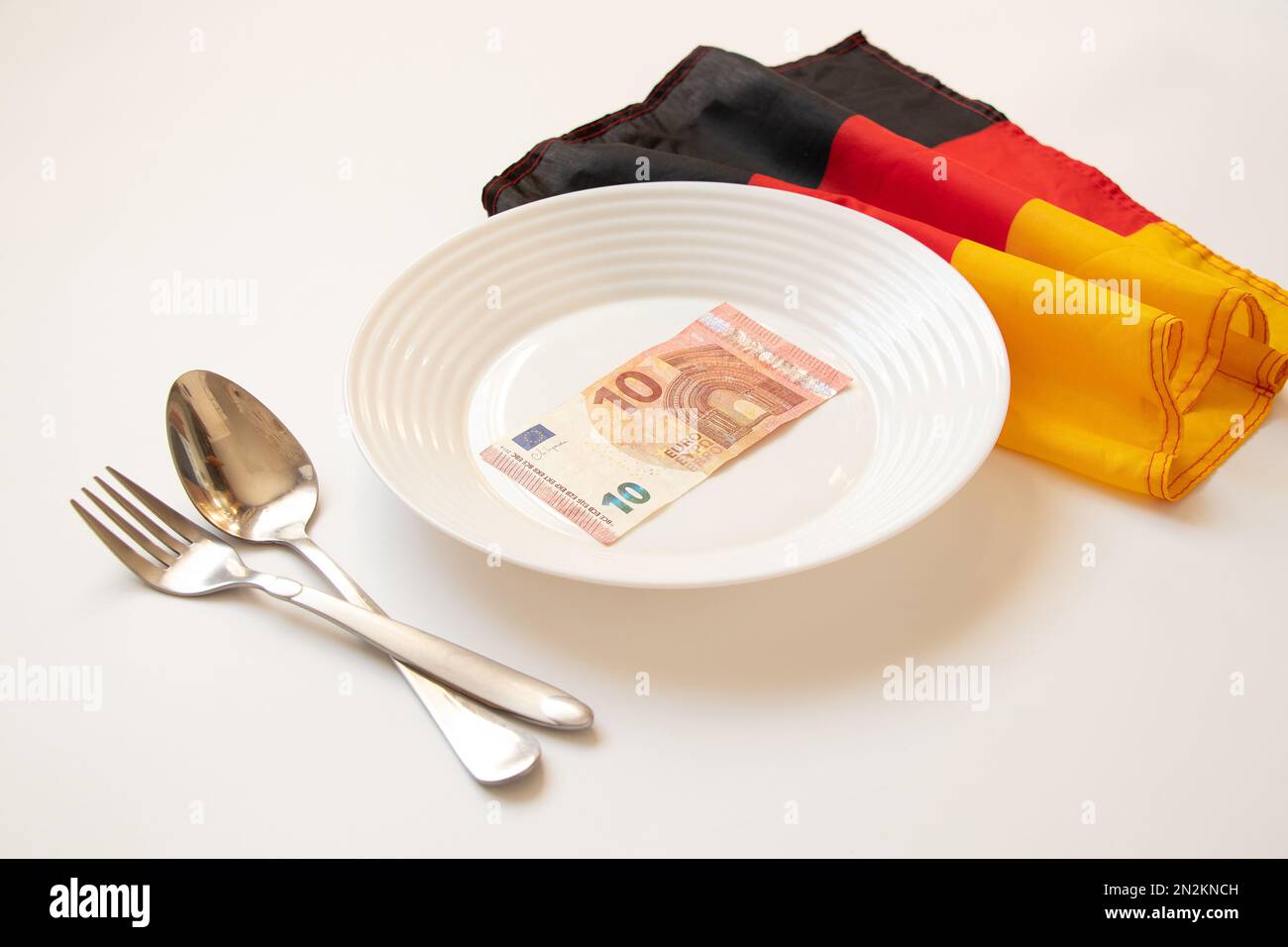 10 Euro liegen auf einem Teller neben einem Löffel und einer Gabel und der Flagge Deutschlands auf weißem Hintergrund, Finanzen und Wirtschaft in Deutschland, Wirtschaft Stockfoto
