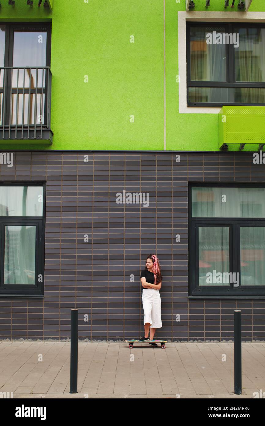 Das junge Mädchen steht mit Schlittschuhlaufen auf der Straße neben dem farbenfrohen grünen Haus. Authentisches Lifestyle-Porträt eines Teenagers Stockfoto