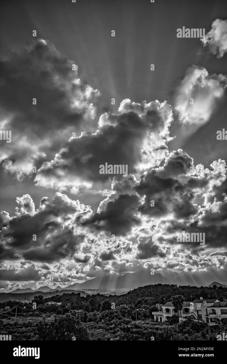 Die Sonne schickt ihre Strahlen an schweren Wolken in dramatischem Himmel über üppigem grünen Tal, umgeben von zerklüfteten Bergen und bestreut mit weißen, von Mauern umgebenen mediterranen Häusern in Spanien, schwarz-weiß Stockfoto