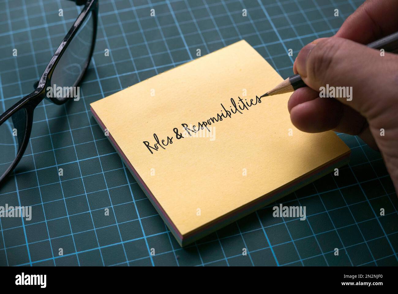 Eine Hand, die Rollen und Verantwortlichkeiten auf einen gelben Memo-Zettel mit einer Brille auf einer grünen Handwerkermatte schreibt. Stockfoto