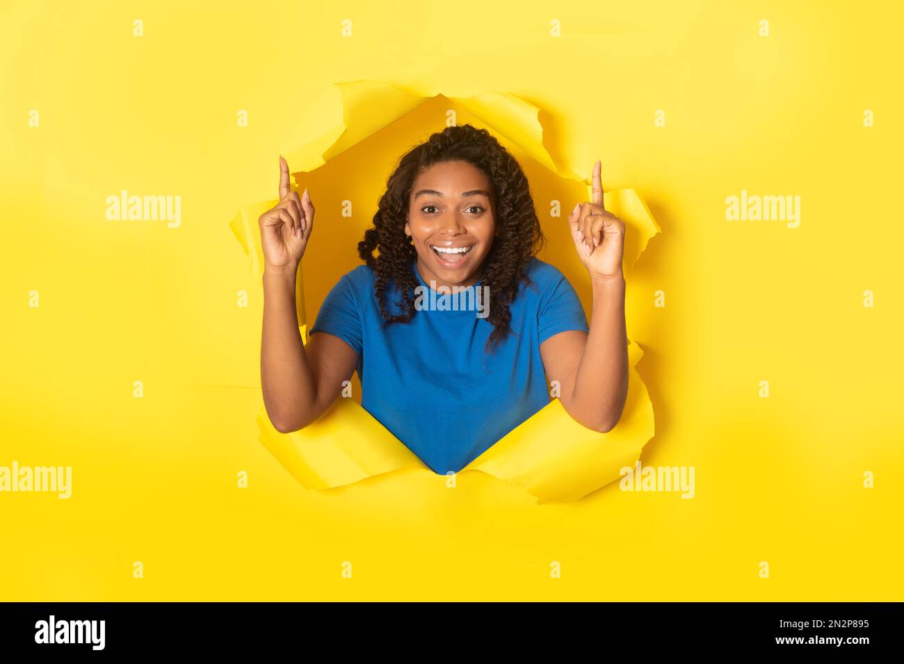 Erstaunte schwarze Dame, die in einem zerrissenen Papierloch mit gelbem Hintergrund posiert, mit den Fingern nach oben zeigt und in die Kamera schaut Stockfoto