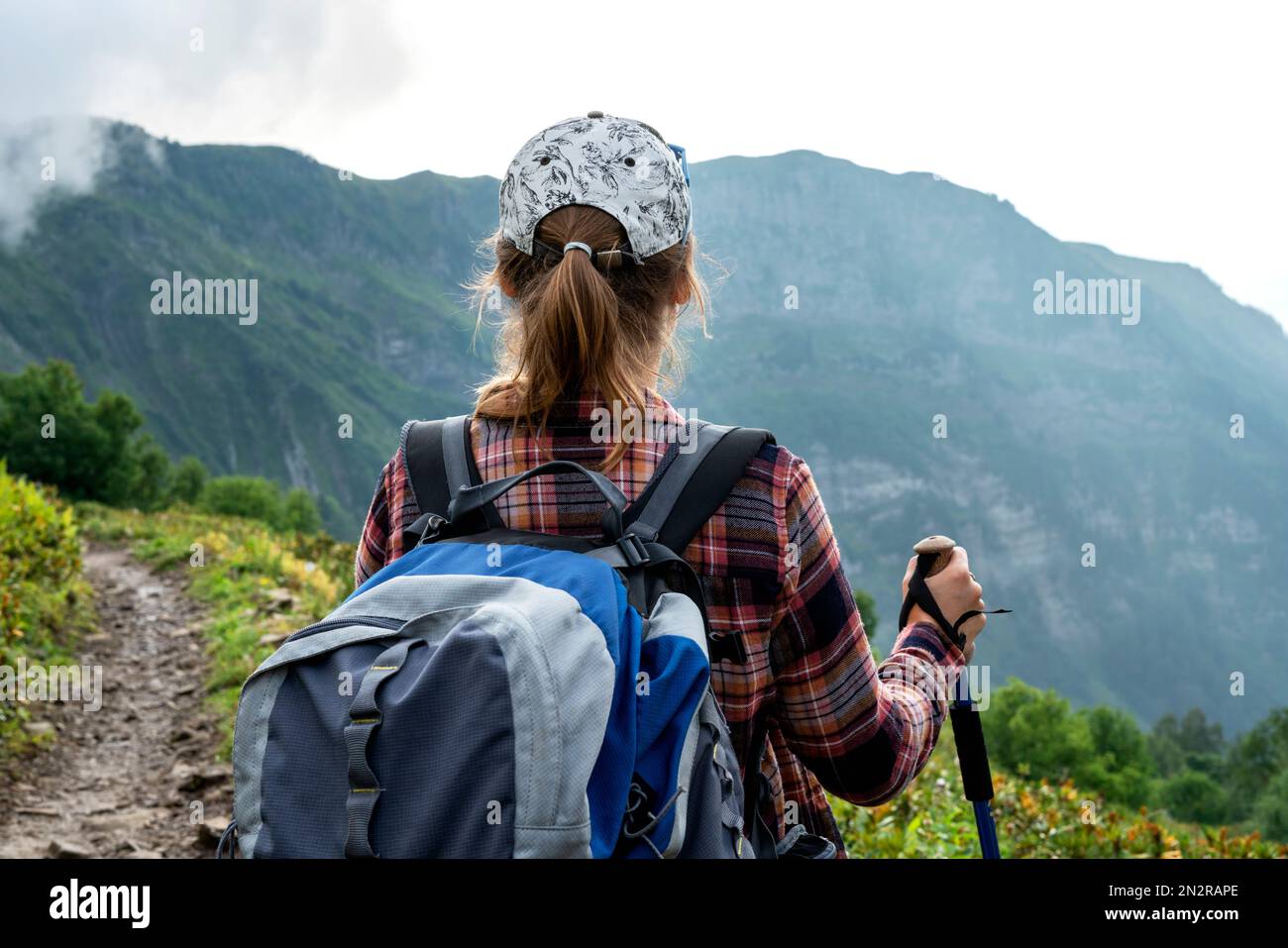 Rückansicht einer jungen Frau in kariertem Hemd und Mütze mit großem Rucksack mit Wanderstöcken, die im Sommer auf dem Bergpfad wandern und gesund und aktiv leben Stockfoto