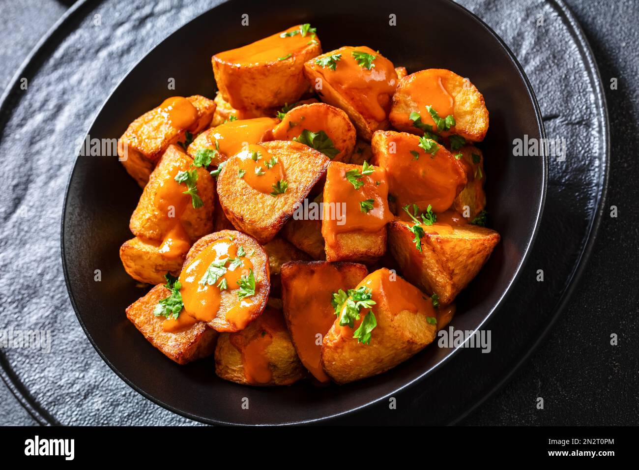 Nahaufnahme von Patatas bravas, frittierten Kartoffeln mit scharfer Sauce in schwarzer Schüssel auf Betontisch, spanische Küche Stockfoto