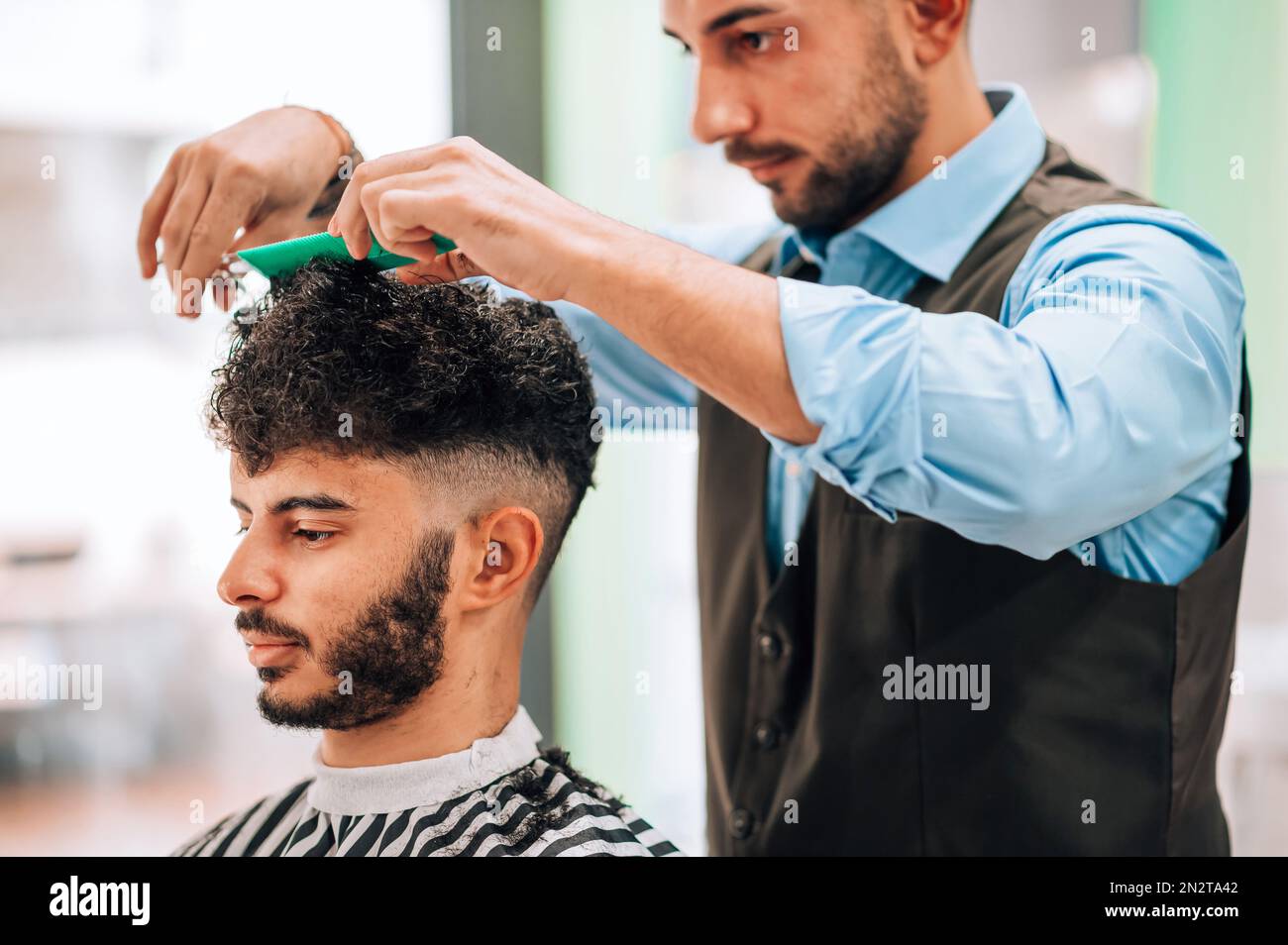 Ein Friseur schneidet lockige dunkle Haare eines bärtigen männlichen Klienten im gestreiften Umhang während der professionellen Frisur im Friseursalon Stockfoto