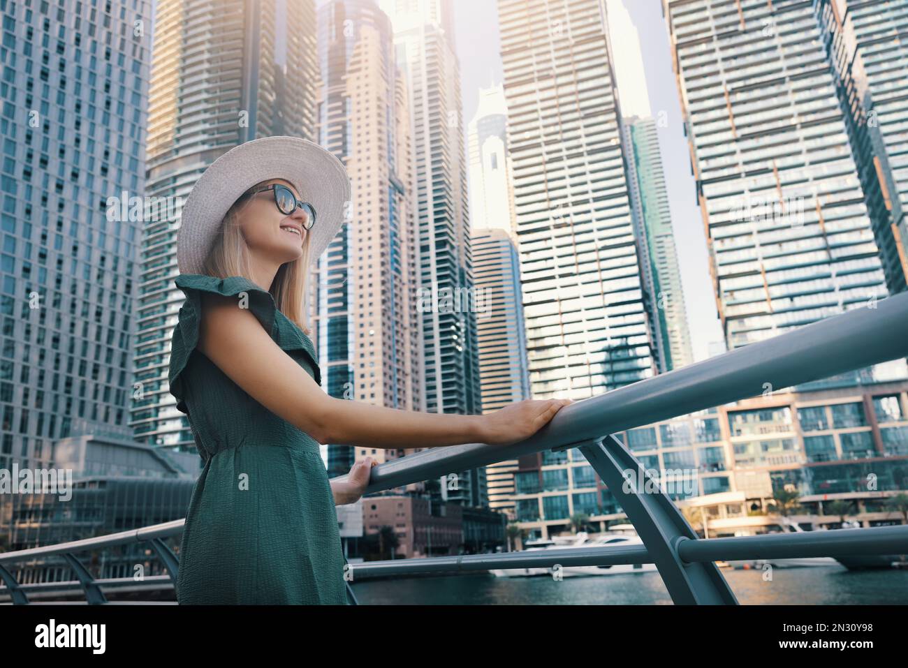 Eine Frau in modischem Sommerkleid und Hut, die den Blick auf den Yachthafen von Dubai mit Wolkenkratzern in den Vereinigten Arabischen Emiraten genießt Stockfoto