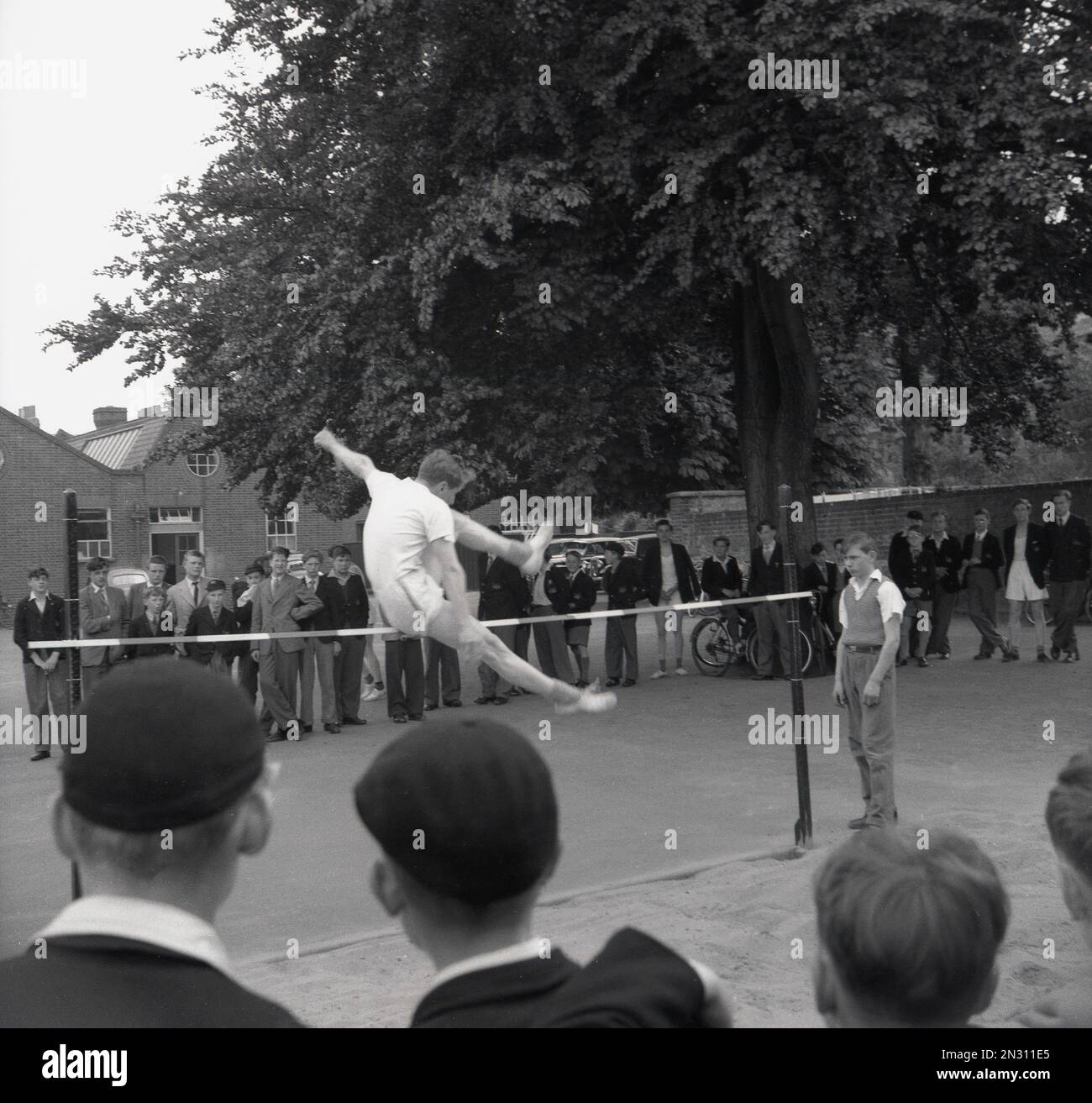 1950er, historisch, draußen auf einem Schulspielplatz, sieht eine Gruppe uniformierter Schuljungen zu, wie ein Schüler in einem High-Jump-Wettbewerb in England, Großbritannien, über die Latte springt. Stockfoto