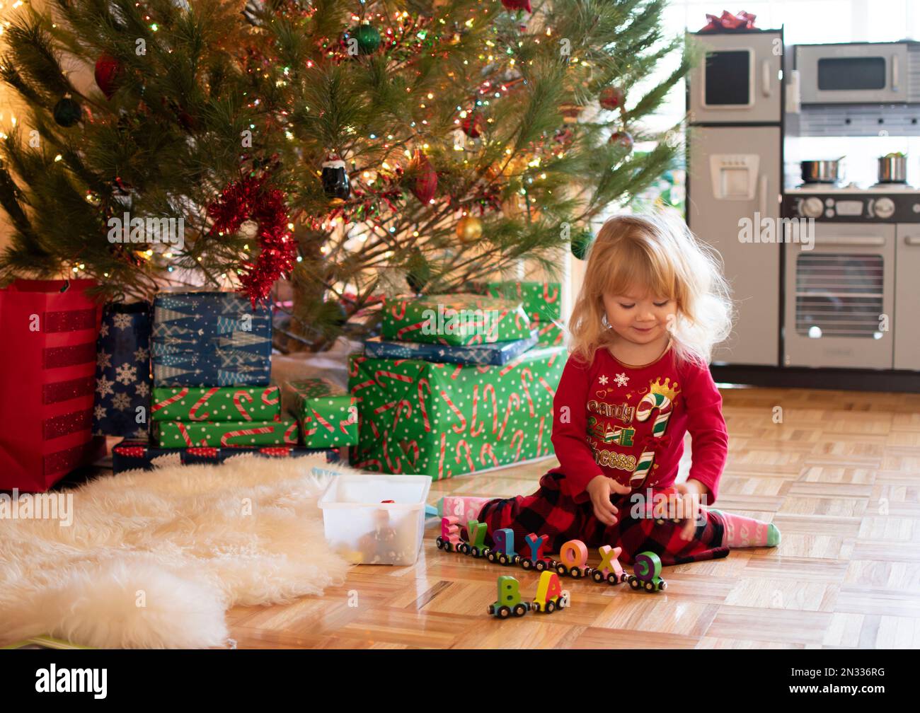 Kleines süßes blondes Kleinkind im Schlafanzug, das an einem Weihnachtsmorgen mit Spielzeug unter dem Weihnachtsbaum spielt Stockfoto
