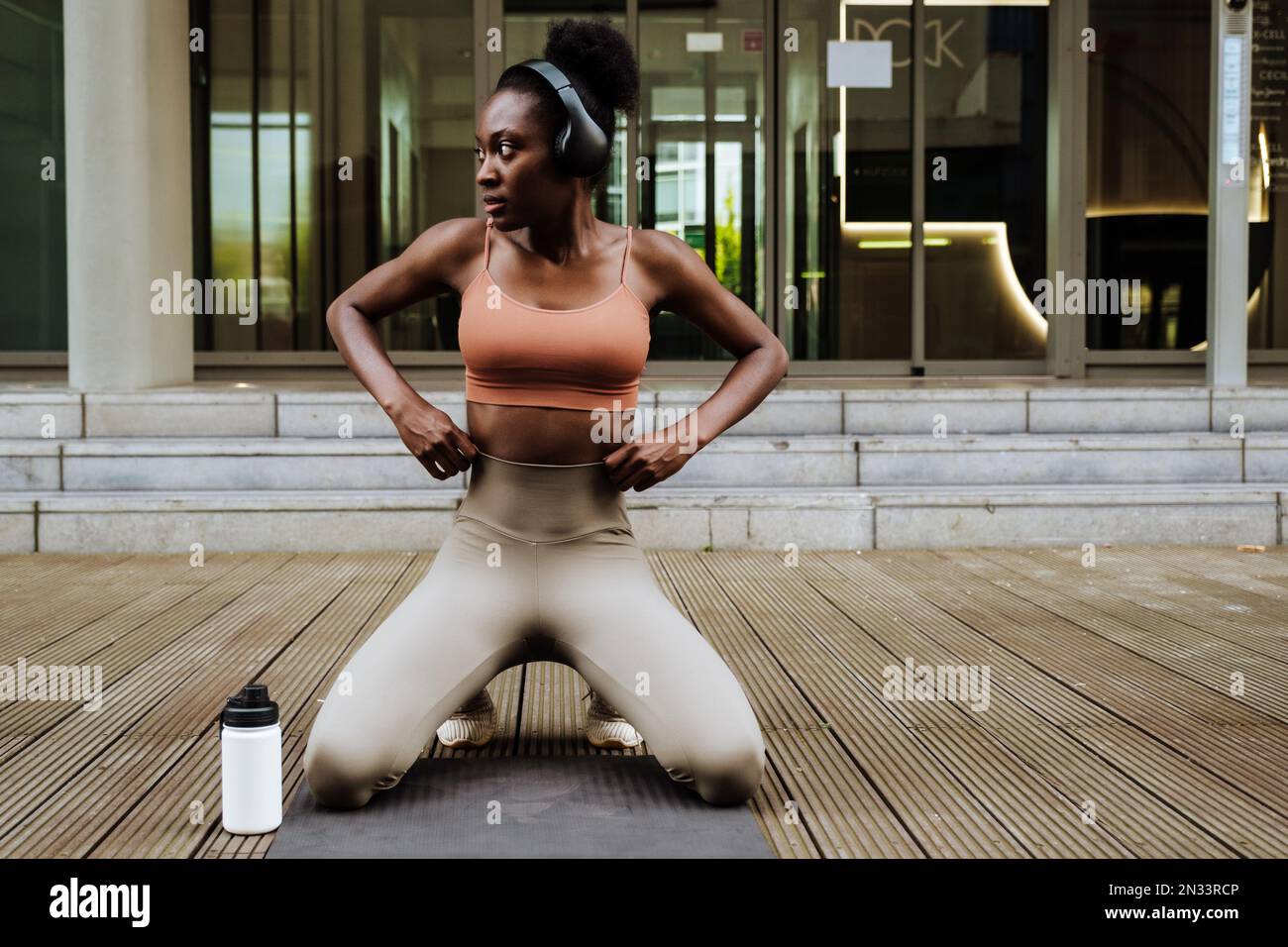 Junge, wunderschöne, ruhige afrikanerin mit Kopfhörern und Trainingsanzug, die auf den Knien auf der Matte steht und zur Seite schaut Stockfoto