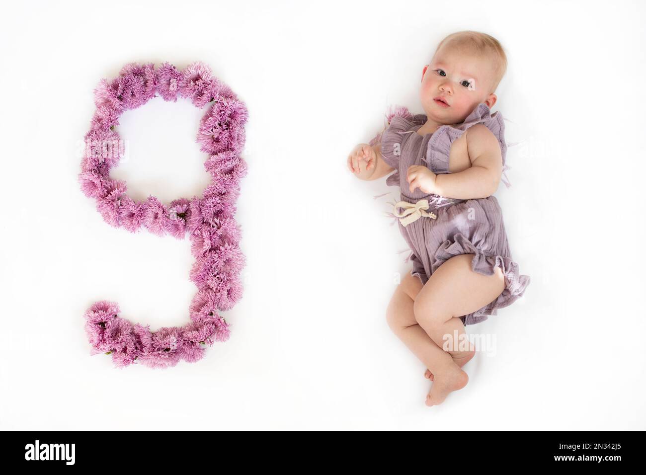 Kleines neugeborenes Mädchen in einem lila Strampelanzug. 9 Monate altes Baby. Neun Monate Baby-Meilenstein. Stockfoto