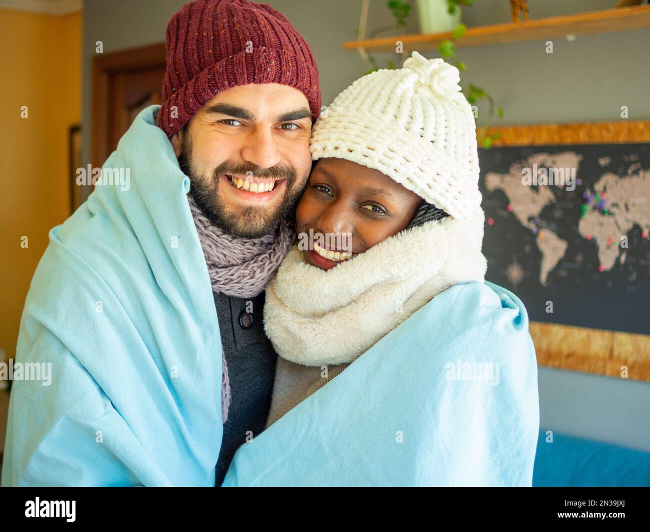 Ein junges Paar, das sich eine Decke teilt, um die kalte Welle zu bekämpfen. Sie tragen Winterkleidung in einem gemütlichen Wohnzimmer. Ich schaue in die Kamera. Stockfoto